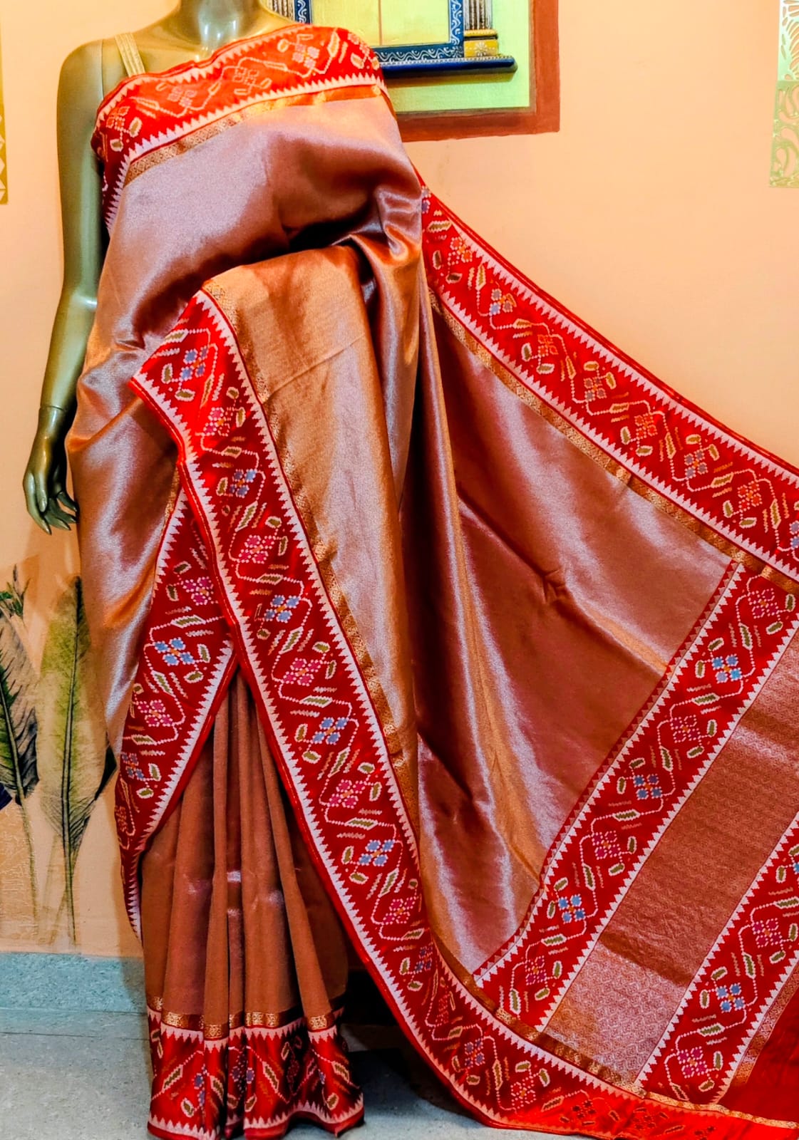 A copper tissue saree draped on the stand, with a wide red patola border in white, blue and green motifs